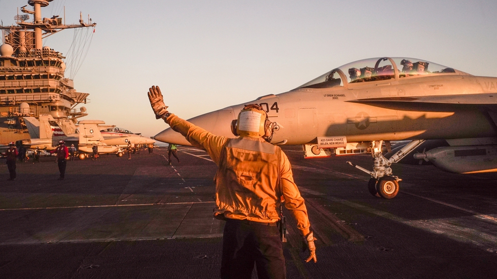 Un operario de cubierta da indicaciones a un F-18 biplaza. De fondo, La Isla, la torreta del portaviones.